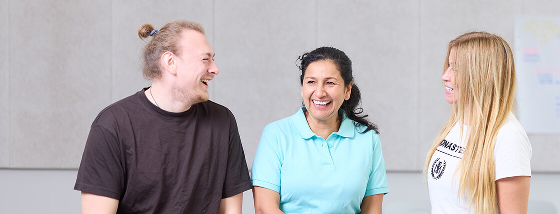 Ein junger Mann und zwei junge Frauen sitzen auf einem Tisch in einer Klasse im Ausbildungszentrum Seegasse und lachen