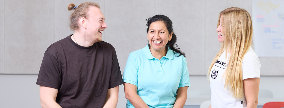 Ein junger Mann und zwei junge Frauen sitzen auf einem Tisch in einer Klasse im Ausbildungszentrum Seegasse und lachen