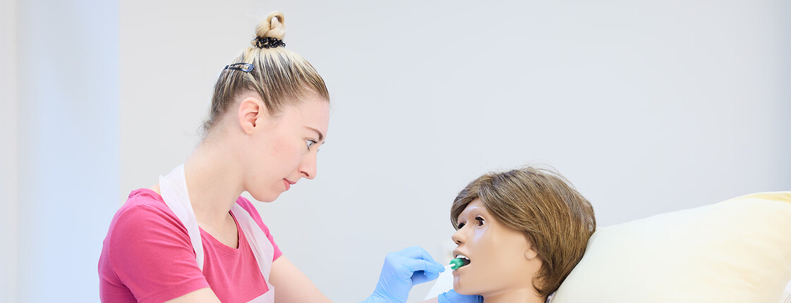 Man sieht eine Trainingssituation, in der eine junge Frau einer Übungs-Puppe im Caritas Ausbildungszentrum Seegasse mit einem Stäbchen den Mund reinigt
